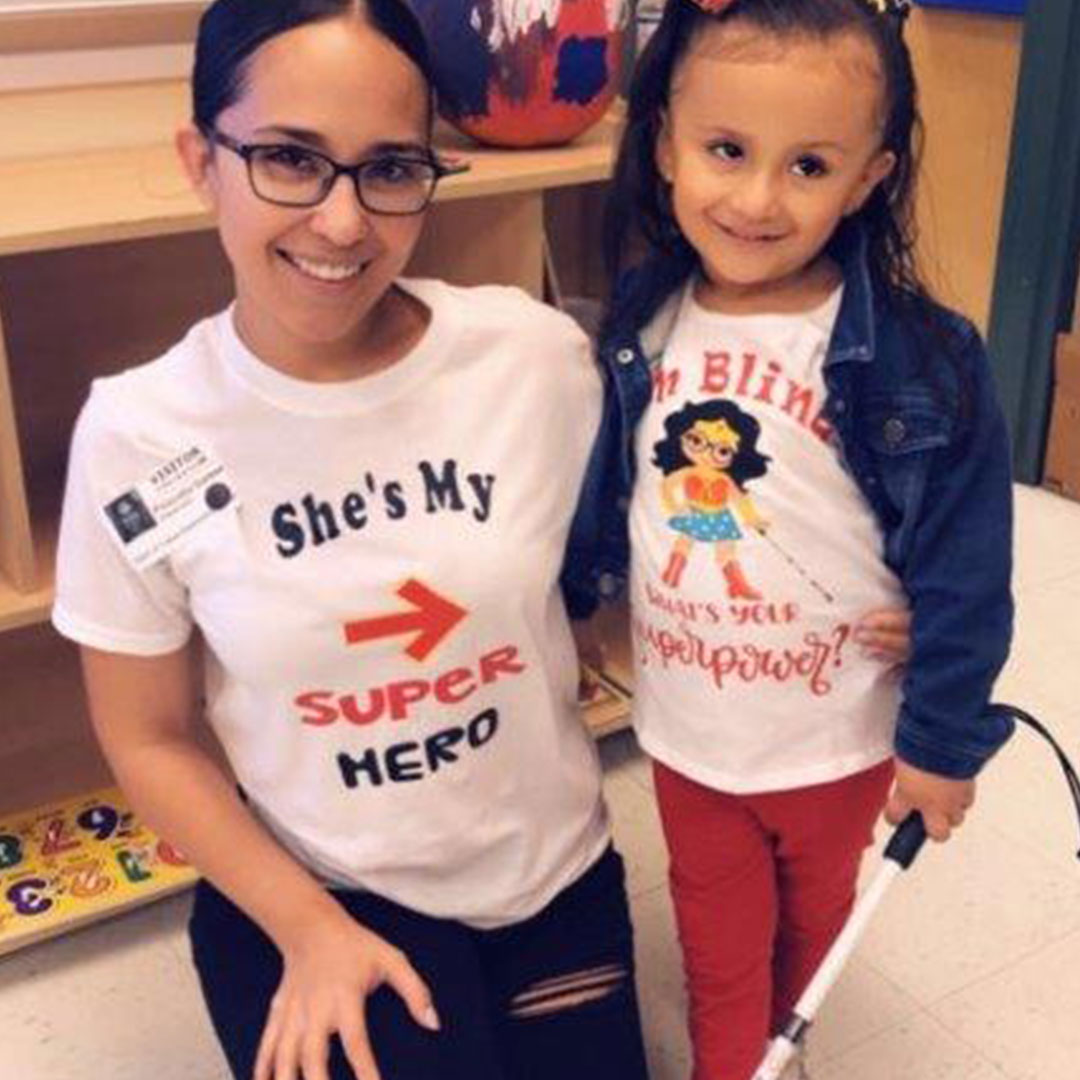 A young girls holds her white cane and wears a Superhero t-shirt.