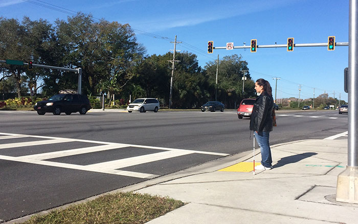A woman with a white cane waits at a crosswalk.