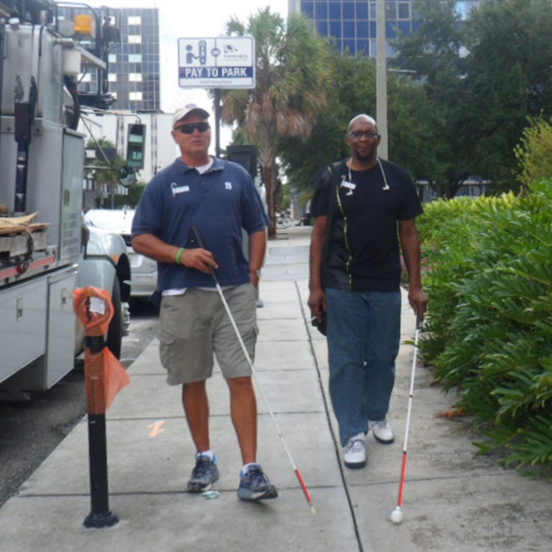 Two men with white canes walk on a city sidewalk.