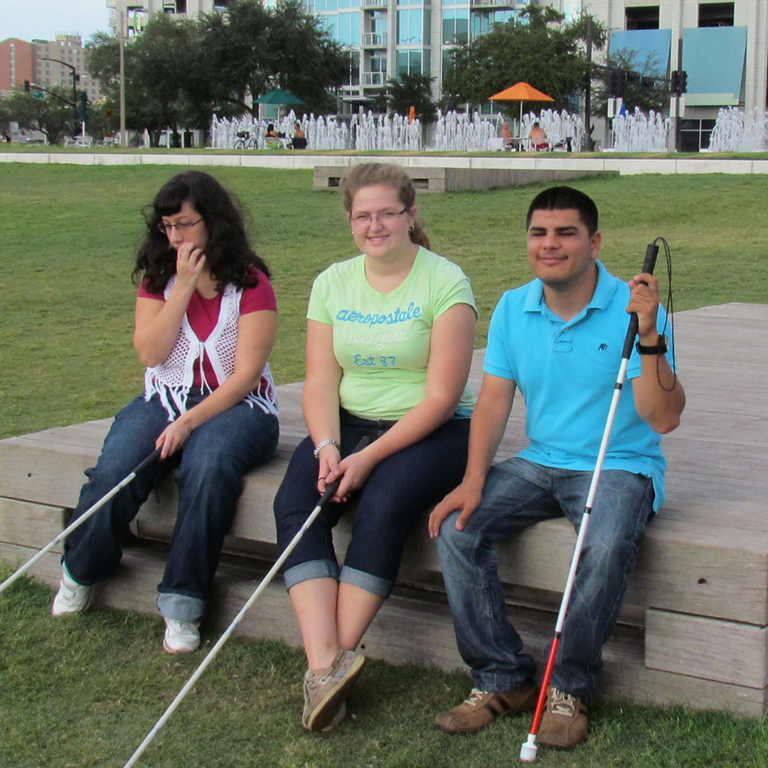 Friends with low vision and white canes rest on a platform in the park.