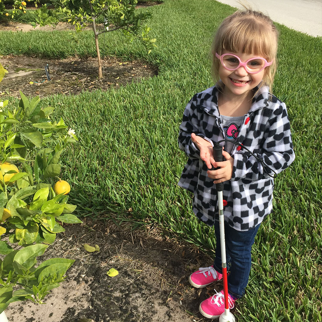 A young girl with low vision holding a white cane walks through a garden.
